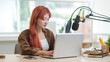 © wattana - Cute young woman with red hair using laptop while recording a podcast in a modern studio.