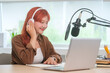 © wattana - Cute young woman with red hair using laptop while recording a podcast in a modern studio.