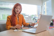 © wattana - Young asian woman graphic designer with red hair is focused on her work, selecting colors for her project at desk.