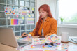 © wattana - Young graphic designer with red hair is focused on her work, selecting colors for her project at desk.