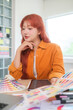 © wattana - Young asian woman graphic designer with red hair is focused on her work, selecting colors for her project at desk.