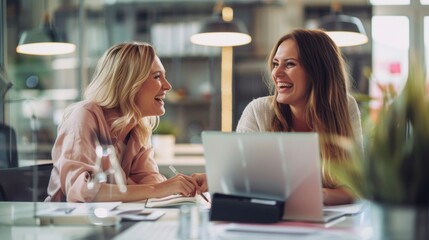 In a contemporary workspace, two women share a moment of pure joy, laughing together as they engage in a lively conversation, surrounded by a bright and inviting atmosphere