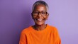 © liliyabatyrova - A cheerful senior black woman beams with joy while wearing stylish glasses and an orange sweater. The vibrant purple backdrop enhances her warm smile