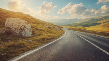 Mountain road scene with rocks, a sunny sky dotted with clouds, and a picturesque asphalt road in the summer evening. Features vintage toning, showcasing a travel background with a highway winding.