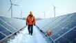 © alihamzatullah - A construction worker walks through a solar field with the solar panels covered in snow. They don’t produce any power like this. Wind turbines for power production are seen at the horizon.