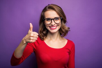 Happy young adult female with glasses and red top gives a thumbs up gesture in front of a purple background, conveying friendliness and approachability.