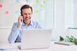 © baranq - Businessman having a phone call while working on a laptop at his desk in a modern office.