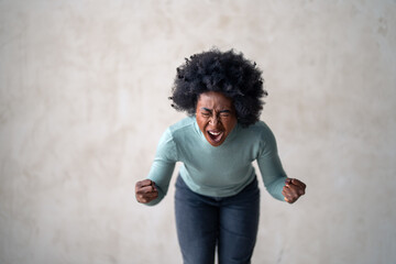 An agitated black female in casual attire stands indoors, fists clenched, with a grey backdrop, embodying frustration and determination.