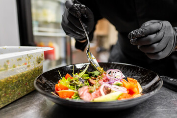 Wall Mural - A close-up of gloved hands garnishing a vibrant vegetable salad with sauce in a black bowl. Perfect for culinary, healthy eating, and food preparation themes.