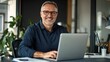© Ganesha - Portrait of happy mid aged business man executive working on laptop in office. Senior company manager investor using computer sitting at desk. Smiling older professional businessman looking at camera