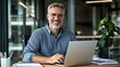 © Ganesha - Portrait of happy mid aged business man executive working on laptop in office. Senior company manager investor using computer sitting at desk. Smiling older professional businessman looking at camera