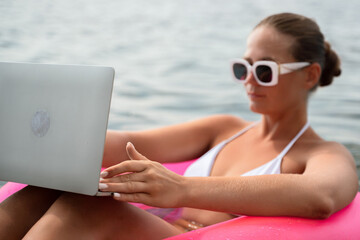 Wall Mural - A woman is sitting in a pink inflatable raft on a lake, holding a laptop. Concept of relaxation and leisure, as the woman enjoys her time in the water while working on her laptop.