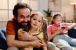 © pressmaster - Father and daughter sharing affectionate moment on couch while another child using smartphone in background creating warm family atmosphere in cozy living room