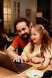 © pressmaster - Father and daughter enjoying time together using laptop in cozy living room setting during evening. Father guiding daughter while she learns computer skills, sitting at table