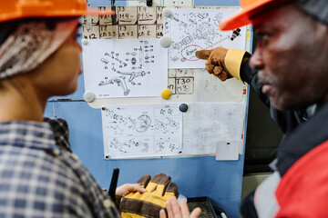 Two engineers reviewing detailed schematics on bulletin board, one pointing at specific part of the drawing while the other listens attentively wearing safety gear