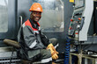 © pressmaster - Worker smiling while operating industrial machinery wearing protective gear Equipment and controls are visible with clear focus on worker's expression and attire
