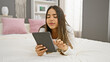 © Krakenimages.com - A young hispanic woman enjoys using a tablet while relaxing on a bed in a cozy bedroom setting.