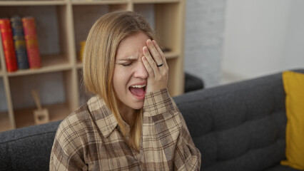 Poster - Blonde woman covering eye with hand, expressing discomfort indoors with a blurred background.