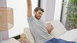 © Krakenimages.com - A relaxed young hispanic man stretching in bed with a coffee mug, representing a cozy morning in a modern bedroom