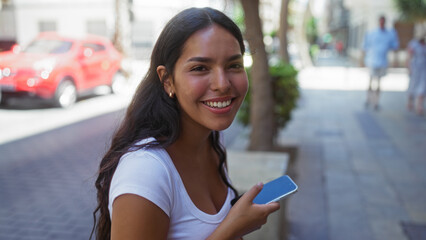 young woman holding a phone and smiling on a city street, exuding happiness and vibrancy in an urban