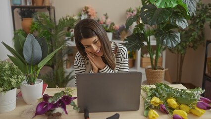 Wall Mural - A cheerful young hispanic woman enjoys a video call at home surrounded by fresh flowers and indoor plants.