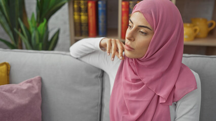 Wall Mural - A thoughtful young woman wearing a pink hijab poses indoors, reflecting tranquility and elegance within a home setting.