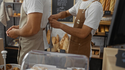 Male bakers working together in an indoor bakery setting, adjusting aprons and preparing for service surrounded by fresh bread and pastries