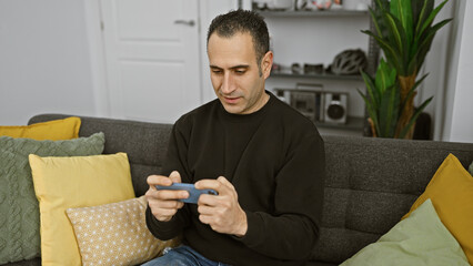 A focused man sits comfortably indoors, using a smartphone with home decor, casual attire, and modern technology.