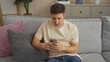 © Krakenimages.com - A young, hispanic, handsome man is sitting on a couch at home, using his smartphone in a modern, cozy living room with stylish decor.