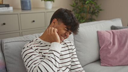 Sticker - A young hispanic man scratching his itchy face while sitting on a couch in a cozy living room, displaying an expression of discomfort.