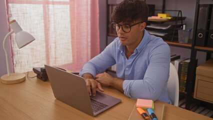 Poster - A young hispanic man working on a laptop indoors in an office setting with shelves and various office supplies around.