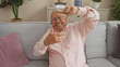 © Krakenimages.com - Elderly man sitting on a couch in a living room making a frame with his hands, wearing glasses and a pink shirt, surrounded by indoor plants and cushions, smiling joyfully.