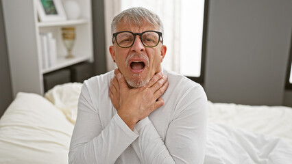 Canvas Print - A gray-haired man in glasses expressing throat pain while seated on a bed in a bedroom setting.