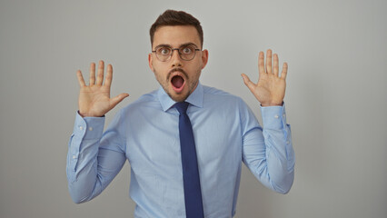 Handsome young hispanic man with a beard and glasses, wearing a blue shirt and tie, posing with a surprised expression, isolated on a white background.