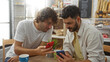 © Krakenimages.com - Men looking at phones together at a bakery, enjoying coffee and pastries, demonstrating communication and technology in a relaxed setting.