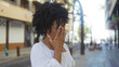 © Krakenimages.com - A beautiful african american woman with curly hair covering her face with her hand while standing on an urban street, depicting an outdoor city scene.