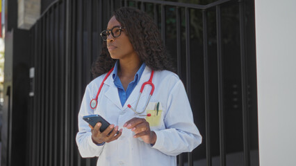 Wall Mural - A beautiful young african american woman doctor with curly hair, wearing a white coat and stethoscope outdoors, looking at her phone on an urban street near a hospital.