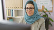© Krakenimages.com - A professional woman wearing a hijab and glasses working at her office desk with a computer, evoking modern diversity.