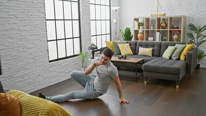 A casual man sitting on the floor in a modern apartment living room, looking tired or pensive.