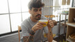 © Krakenimages.com - Young hispanic man with beard and tattoos drinking coffee in a cozy bakery with pastries in the background.