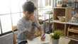 © Krakenimages.com - Young, hispanic, man with a beard and tattoos sits pensively in a bakery, writing in a notebook with shelves of bread and pastries in the background