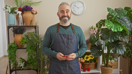 Wall Mural - A smiling bearded man in an apron stands in a flower shop filled with various plants, portraying a friendly florist.