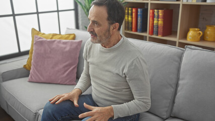 Poster - Middle-aged bearded man sitting thoughtfully on a couch in a cozy living room, conveying a sense of contemplation and comfort.