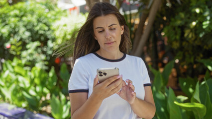 Sticker - Young woman using smartphone outdoors in city park during sunny day with lush green foliage in background