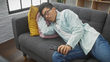 A young asian man sleeps on a couch with colorful pillows in a cozy apartment living room, remote in hand.