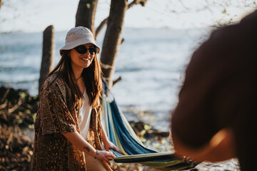 Poster - A woman in sunglasses smiles while setting up a hammock with a friend beside a serene lake surrounded by trees.