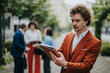 © qunica.com - A businessman in an orange suit reviews documents outdoors while his colleagues engage in a discussion in the background.