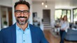 © Pinklife - Smiling man in blue blazer and light blue shirt standing in a bright, modern office environment, with colleagues and a well-decorated room in the background.