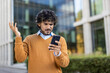 © Liubomir - Man with curly hair standing outside modern office building looks at phone with frustration. Wearing a brown sweater, he expresses confusion or dissatisfaction with technology issues.