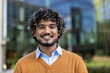 © Liubomir - Smiling young man with curly hair standing outdoors. Wearing casual sweater and shirt in front of modern glass building. Captures friendly, approachable urban city vibe with focus on confidence